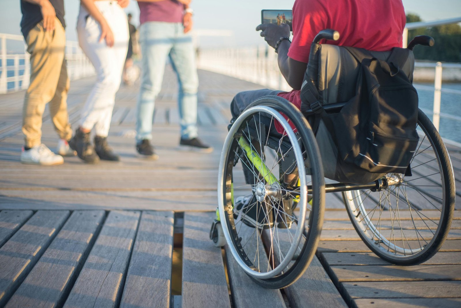 A wheelchair user converses with friends outdoors on a sunny boardwalk.